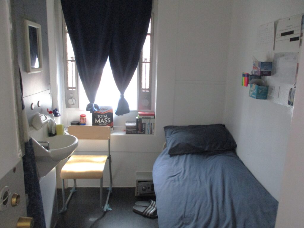 Photograph of a single cel. A bed is on one side, with a chair and sink on the other side. The occupant's personal effects are evident around the room.