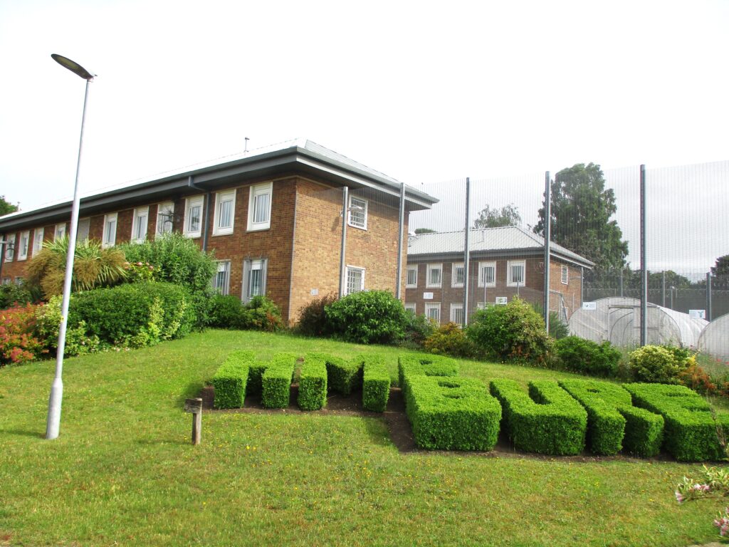 Photograph of the exterior of HMP Bure, with buildings in the background. In the foreground can be seen a box hedge with the name of the institution cut into it.