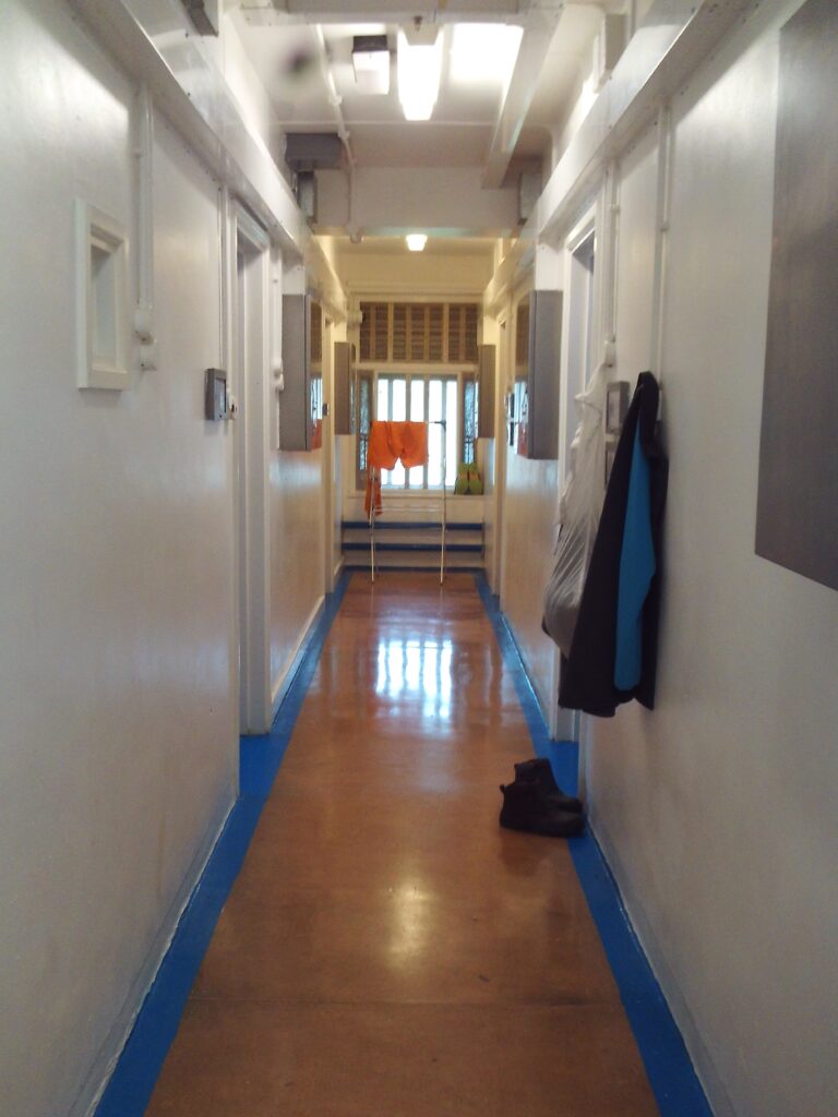 Photograph of a corridor leading onto A to F wings. Light from a window at the far end reflects off the laminate flooring and doorways can be seen on either side of the corridor.
