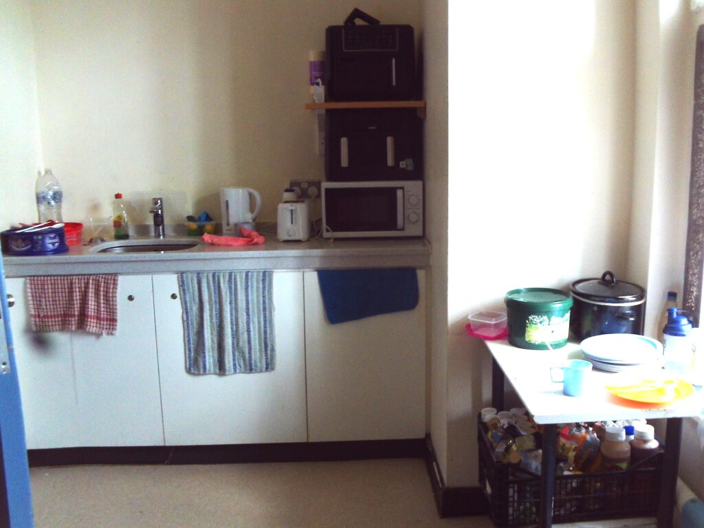 Photograph of the A to F wing kitchen. Basic facilities including microwaves, a toaster, a kettle and a sink can be seen, as well as pots and pans and ingredients.