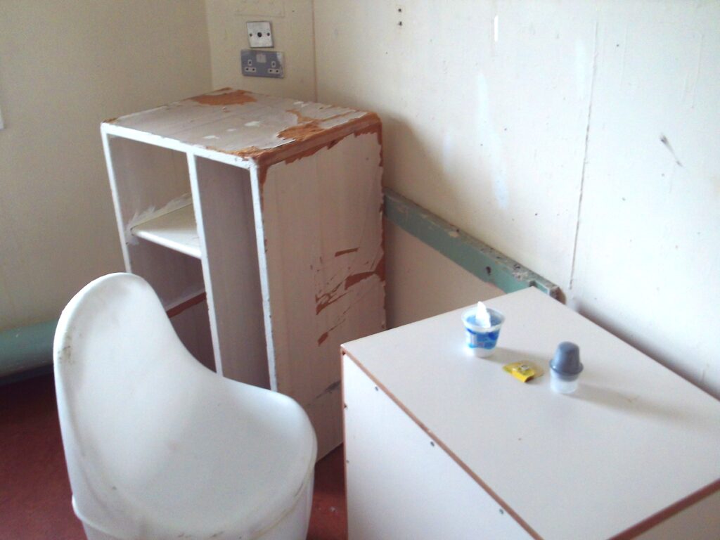Photograph of a first night cell in which can be seen some damaged items of furniture and some litter left by the previous occupier.