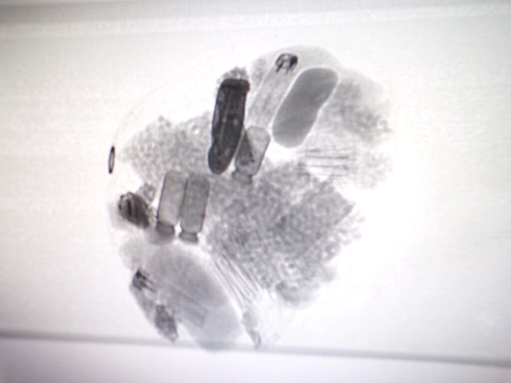 A photograph of an x-ray scan of a package intercepted by officers - drone package recovered in June 2026. Includes bags of tablets, packets of organic material (suspected to be cannabis), and a folding knife (top left of image).