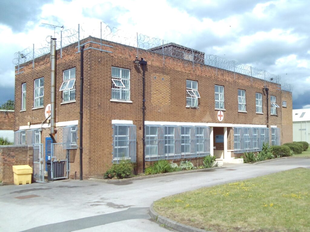 Photograph of the exterior of the health care building, a two-storey brick structure. The windows on the ground floor have metal grilles on them. There is a path and a small area of grass outside the front of the building.