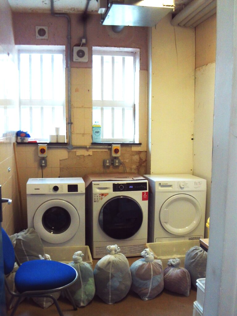 Photograph of a room in which three washing machines  can be seen.  In front of the machines are several bags of laundry and a chair.