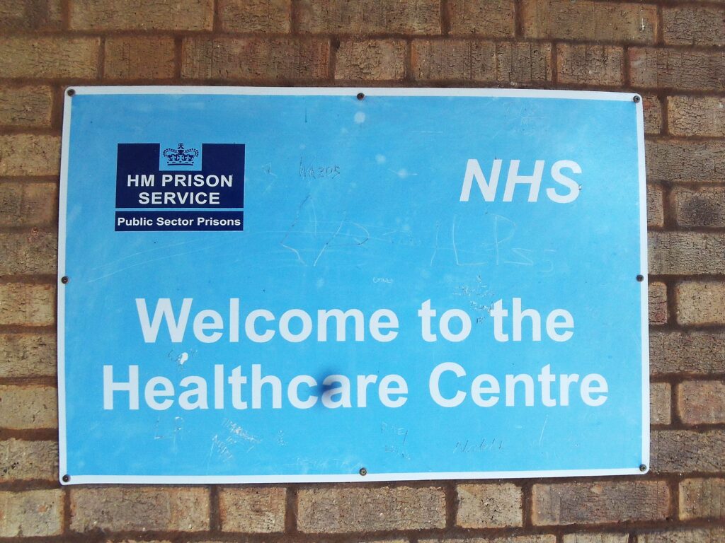 Photograph of the sign welcoming visitors to the health care centre. the blue sign has HMPS and NHS logos on it.