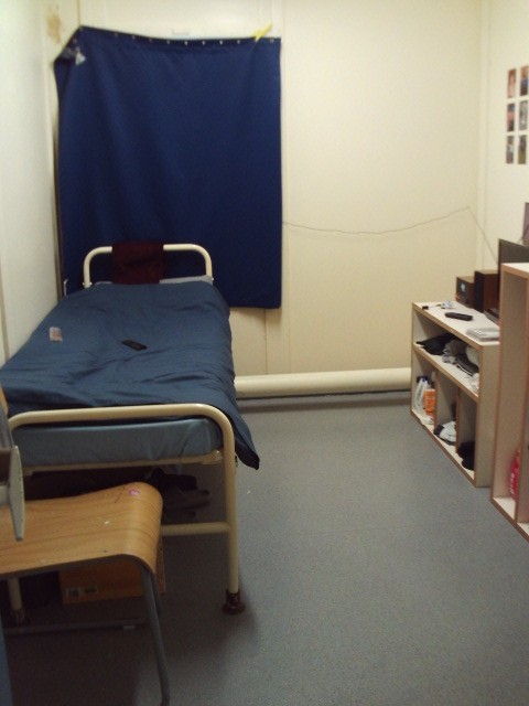 Photograph of an A wing cell. A bed and chair can be seen on one side, with a desk and other furniture arranged against the opposite wall.