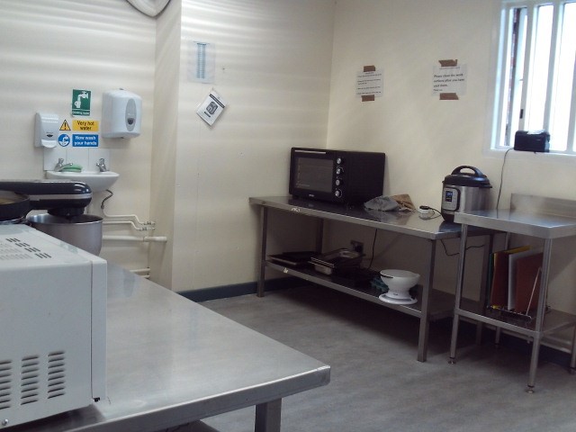 Photograph of a communal; kitchen on G wing south, the semi independent living unit.. Basic catering facilities, including microwaves, and an air fryer are arranged on and underneath stainless steel kitchen units.