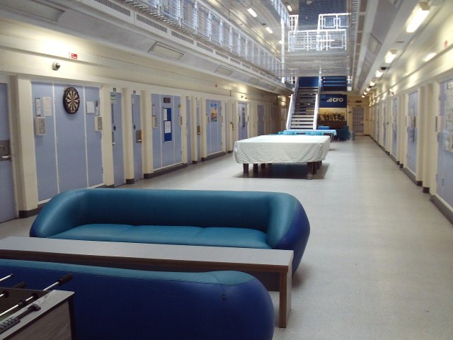 Photograph of the Association area on D wing. There are some sofas arranged around a long, thin coffee table, some pool tables and a darts board. Cell doors can be seen on either side of the space and a staircase leads up to a landing.