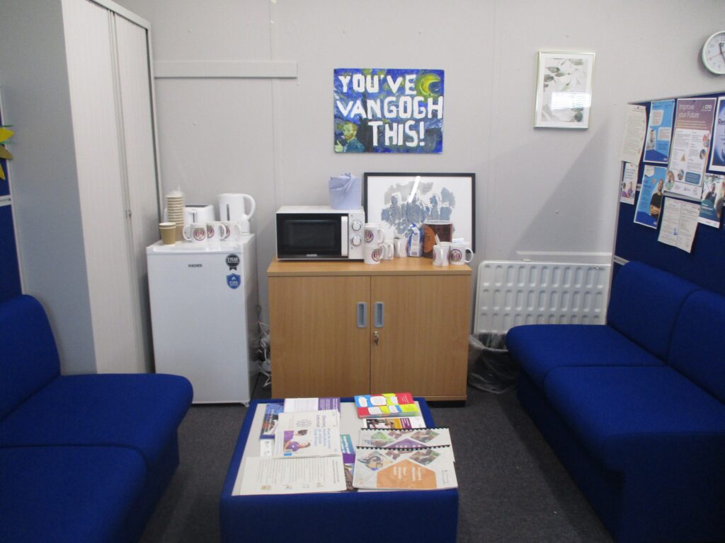 Photograph of the resettlement hub, showing some seats around a table on which there is some literature. Furniture including a storage cabinet, and a fridge, microwave and kettle are behind the seats.