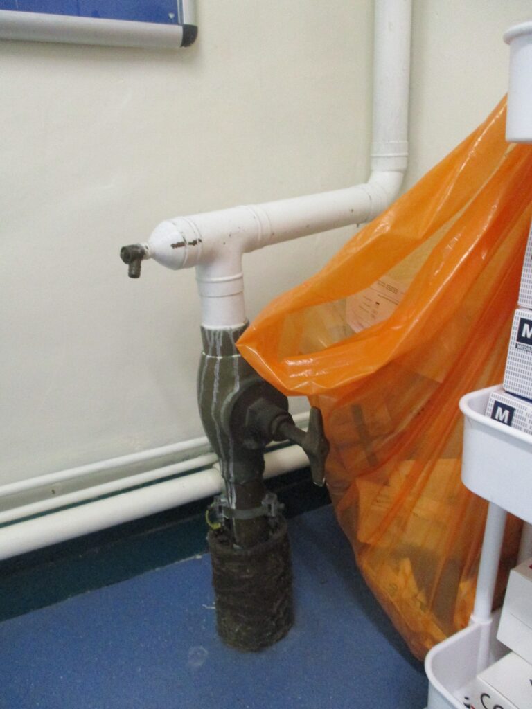 Photograph of an open rubbish bag and some patched up, damaged pipes in a clinical room.
