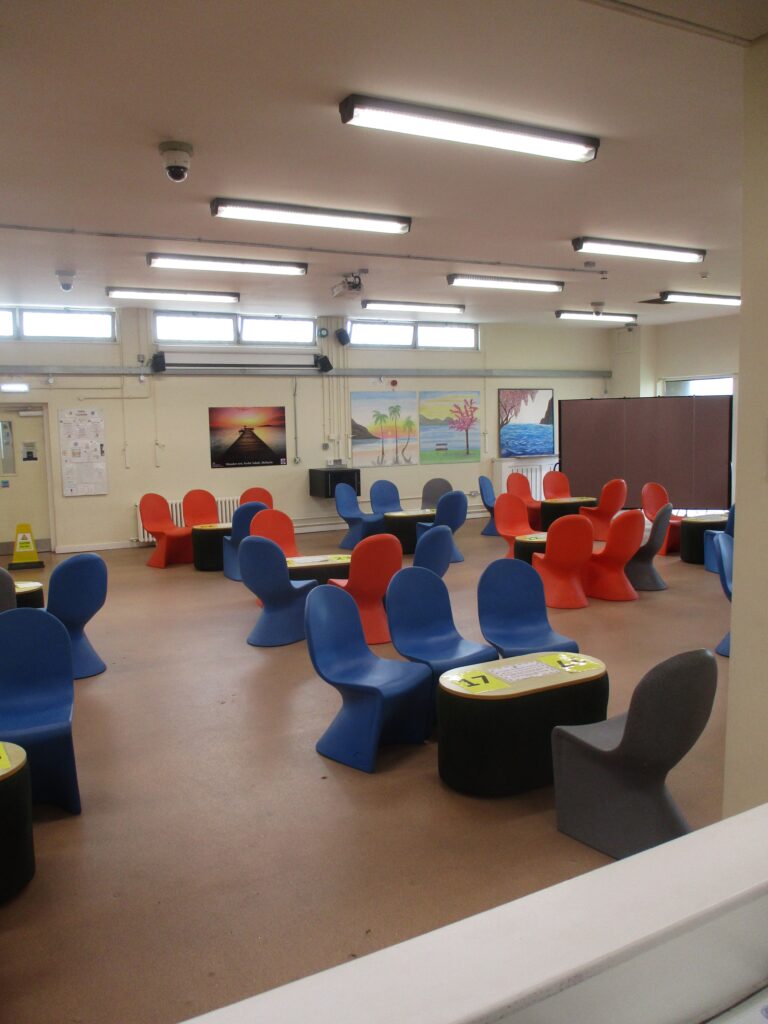 Photograph of the visits hall, showing groups of chairs arranged around low tables. There are posters and art on the back wall and there is a large screen in one corner.