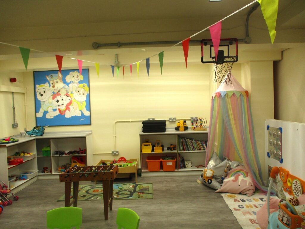 Photograph of children's rea in the visits hall, with play equipment arranged on shelving units and on the floor around the edge of the room.