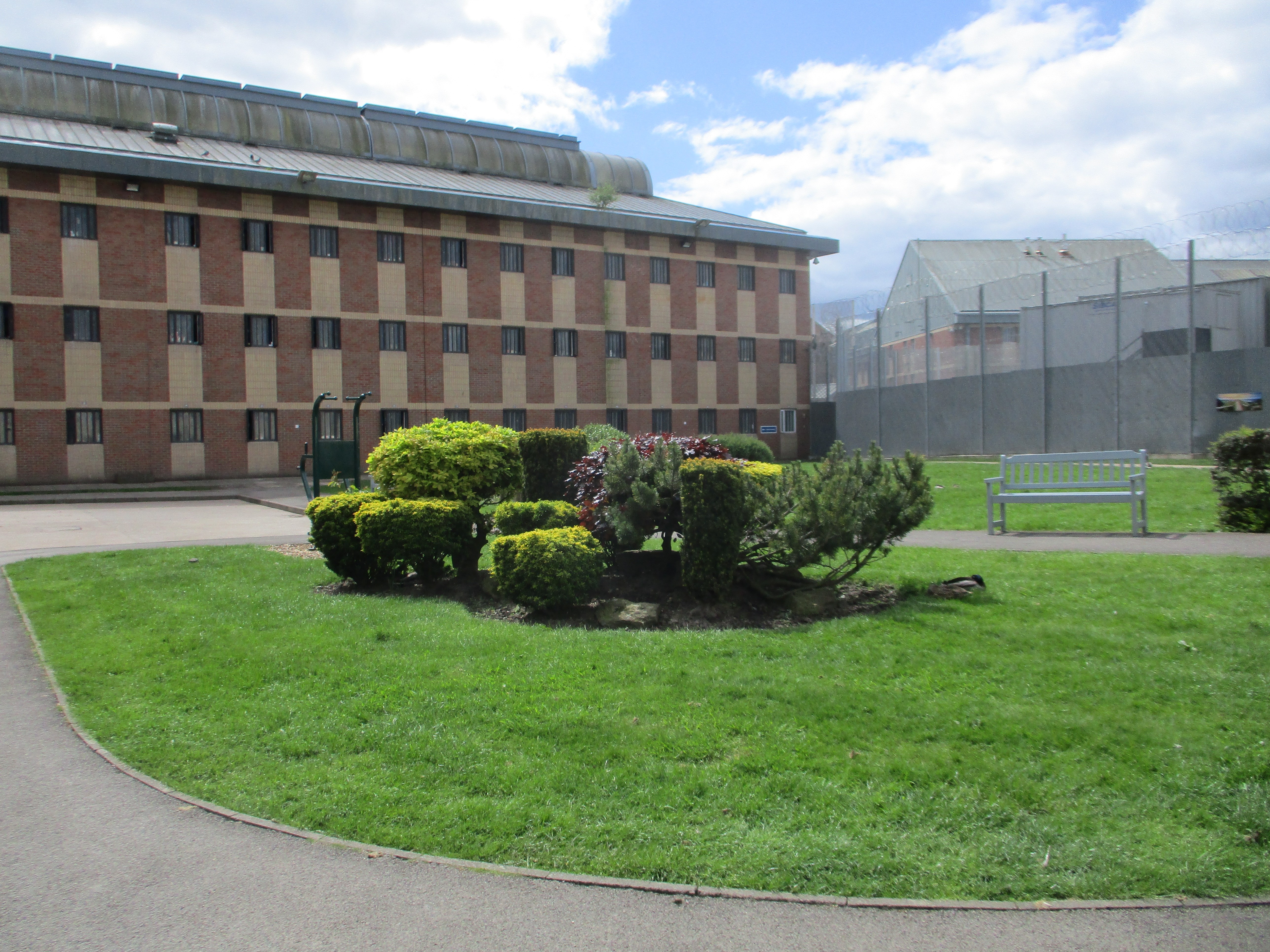 Photograph of the exterior of HMP Risley, with gardens in the foreground and a five storey brick building to te rear. A high fence can be seen to the right, with more buildings behind it.