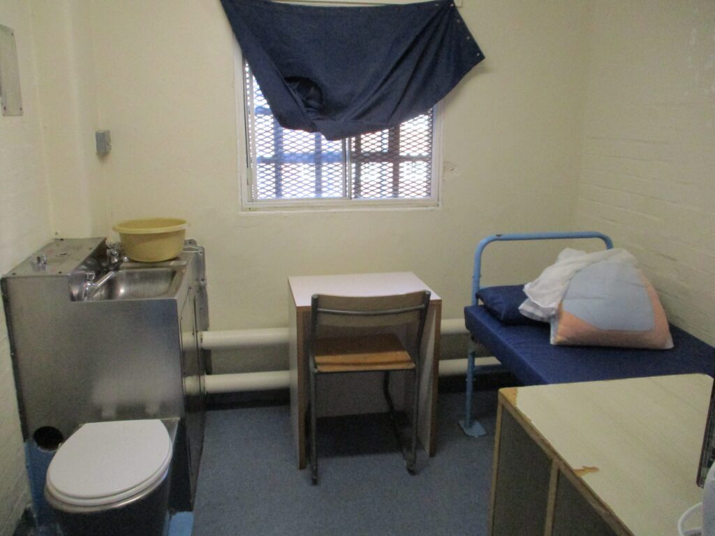 Photograph of a first night cell. There is a sink and toilet on one side, a chair and small desk in the middle, and a bed to the other side.