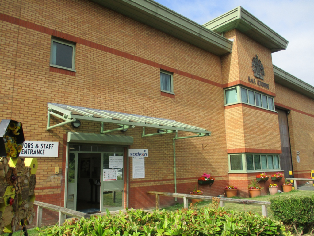 Photograph of a brick building with a sign saying "Visitors & Staff Entrance" and a glass door under a metal awning. There are flower pots and greenery along the walkway.