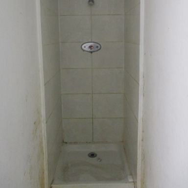 Photograph of a small, narrow shower stall with white tiled walls and a white shower base. The shower features a single control knob and a showerhead positioned high on the wall, with visible dirt or discoloration on the lower walls.