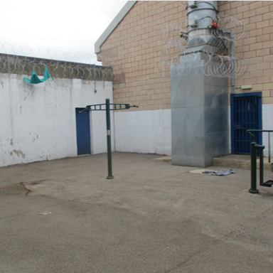 Photograph of an outdoor prison exercise yard enclosed by high walls topped with barbed wire and wire coils. The yard contains metal exercise equipment, including pull-up bars and a stepper machine, with a secured blue door and ventilation duct visible on a brick building wall.