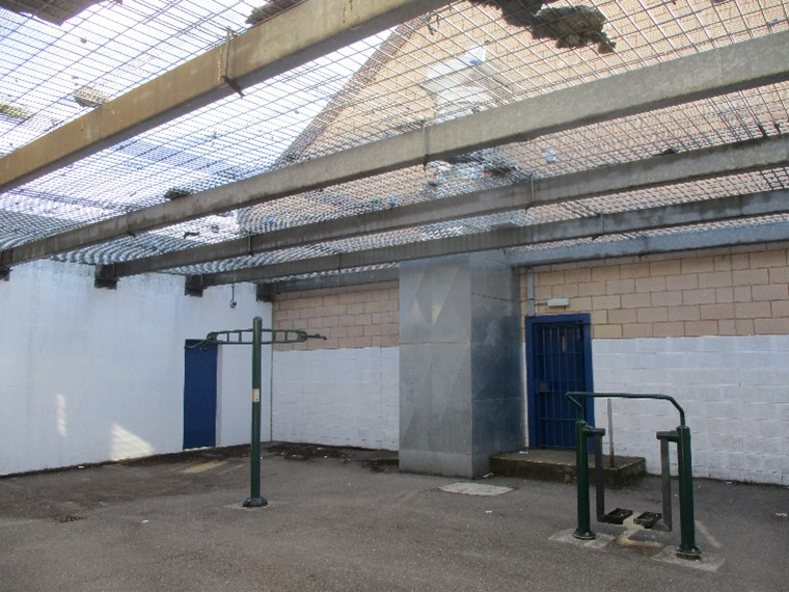 Photograph of an outdoor exercise area enclosed by high walls and a metal mesh ceiling, featuring two green metal workout stations on a concrete floor. The space includes a blue door, a large metal ventilation duct, and a partially painted white and beige brick wall.