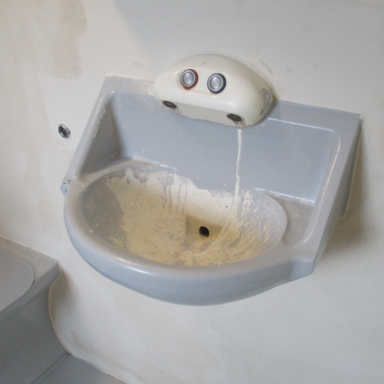 Photograph of a wall-mounted sink, showing signs of wear and discoloration, with two push buttons on top for water control.