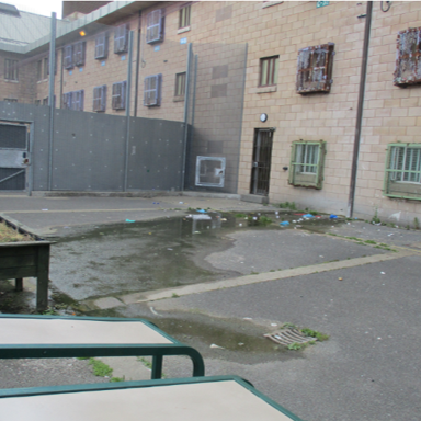 Photograph of an outdoor prison yard enclosed by high brick walls with barred windows and a tall metal fence topped with barbed wire. The yard contains two empty benches, a raised planter box, scattered debris, and a large puddle on the concrete ground.