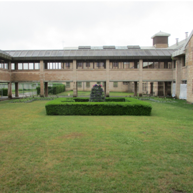 Photograph of a courtyard enclosed by a two-story building with brick walls and numerous windows. The courtyard features a well-maintained lawn and a square hedge centrepiece.