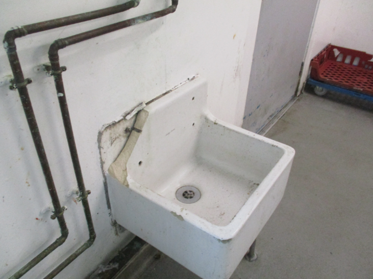 Photograph of an old, wall-mounted broken utility sink with visible wear and chipping paint around edges and mounting area. Rusty metal pipes run along adjacent wall, and a red plastic crate is partially visible on a grey floor nearby.
