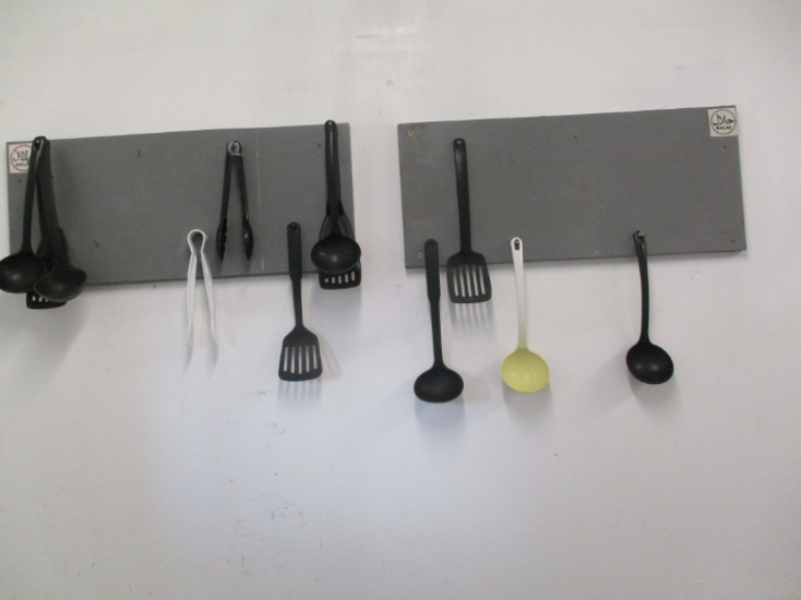 Photograph of two grey wall-mounted racks holding various kitchen utensils, including ladles, spatulas, and tongs. Utensils are mostly black with one white and one yellow ladle, arranged with some hooks empty.
