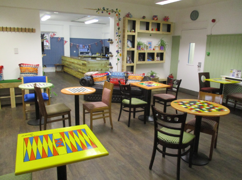 Photograph of the 'enrichment hub' featuring multiple small tables with colourful board game patterns, including backgammon and checkers, surrounded by various wooden chairs. The room has a wooden floor, decorative cushions, wall shelves with plants and books, and a counter area in the background.