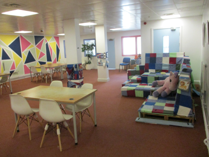 Photograph of the sensory room in the visits hall. The room is colourful and spacious featuring tables with white chairs, a patchwork sofa with a stuffed unicorn toy, and a vibrant geometric mural on one wall. The room has a warm, inviting atmosphere with natural light from windows.