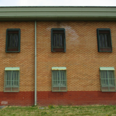 Photograph of a brick building featuring six windows with metal bars. A vertical pipe runs down the centre of the wall.