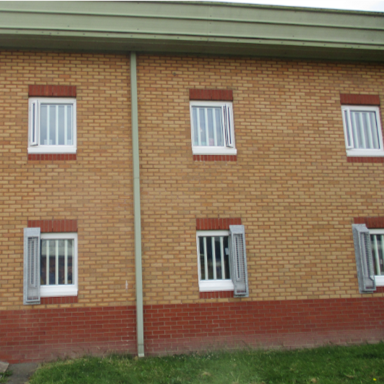 Photograph of a brick building featuring six barred windows arranged in two rows. A vertical drainpipe runs down the centre of the wall, with a grassy area in front.