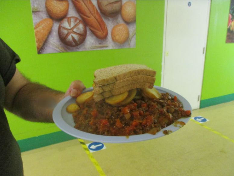 Photograph showing a hand holding a plate with a serving of a mince-based meal with sliced potatoes and a stack of sliced bread. Background features a green wall with a poster of various breads and a white door.