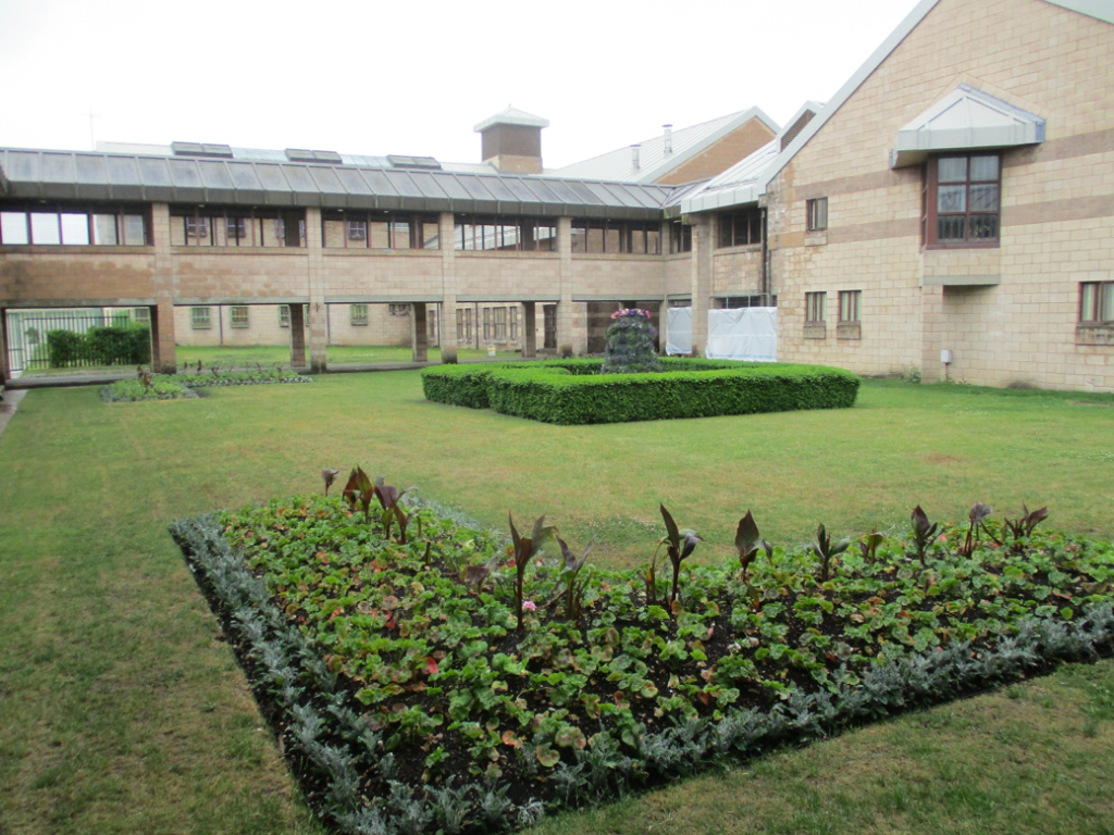 Photograph of a courtyard garden surrounded by beige brick buildings with large windows and a covered walkway. The garden features neatly trimmed hedges and flower beds with various plants.