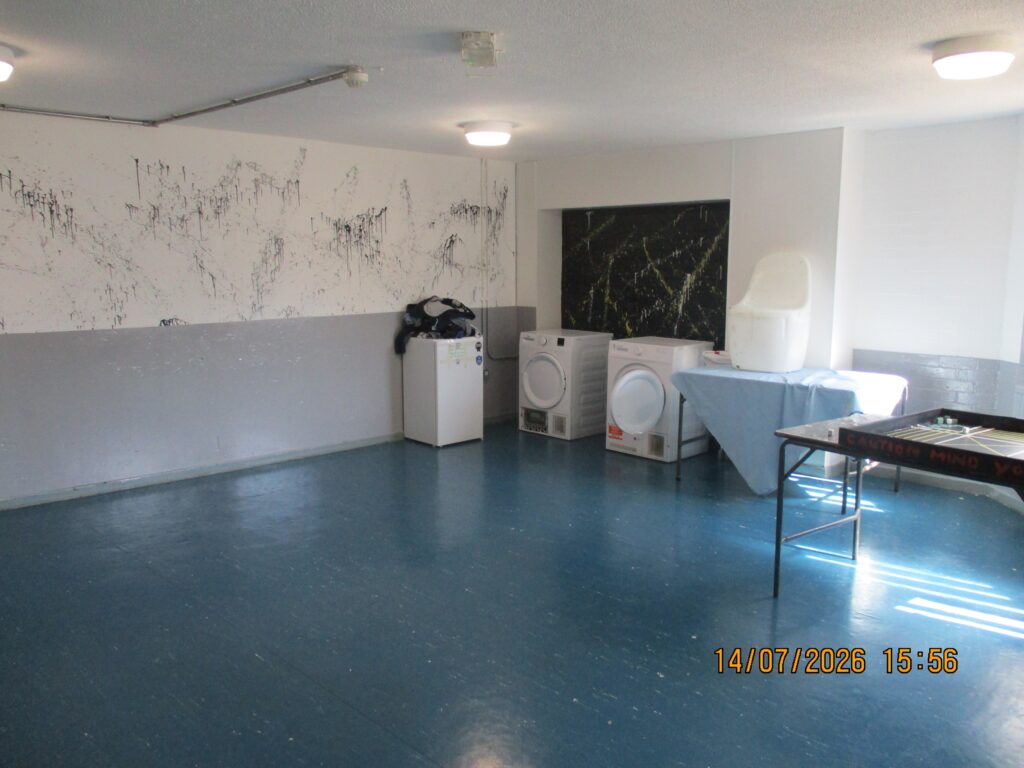 Photograph of an association room. The walls are painted grey and white and there are washing machines and a table to one side.