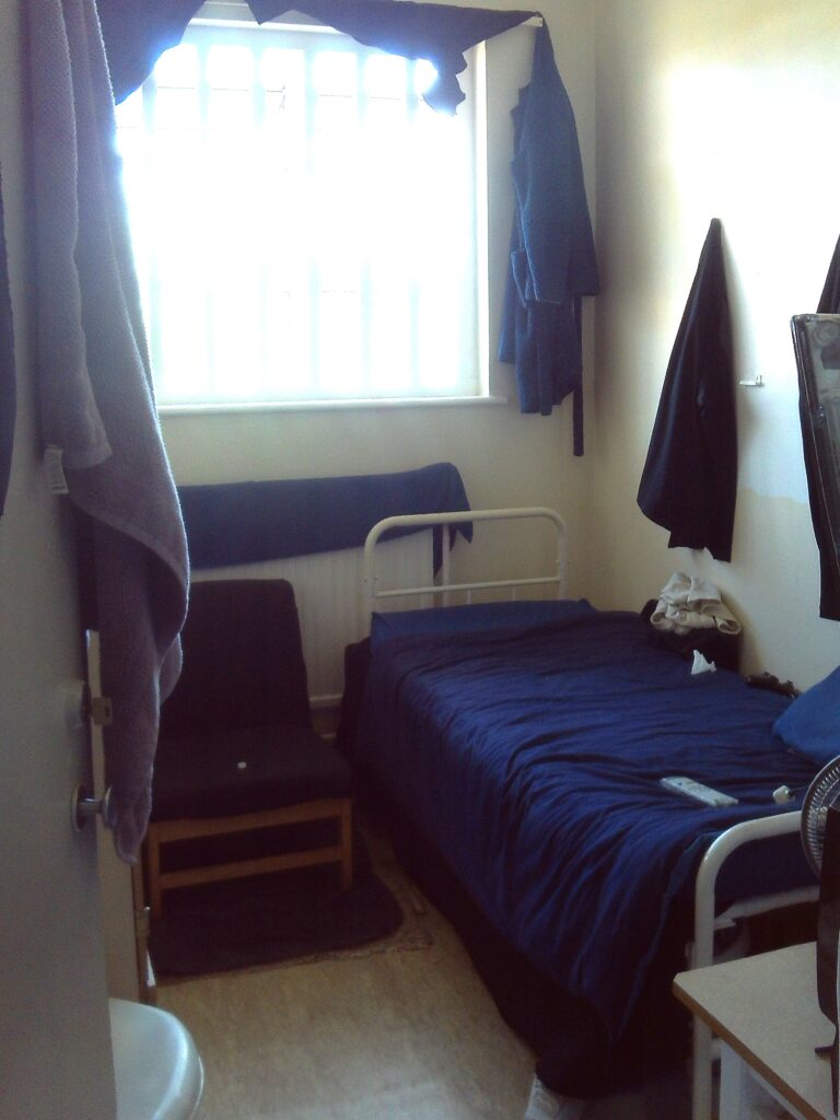 Photograph of a typical cell. It is clean and tidy. There is a bed to one side, a chair next to it, and further items of furniture.