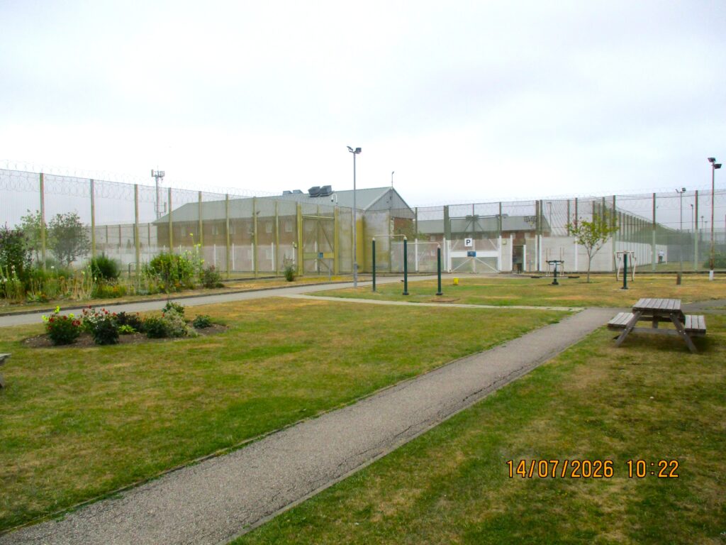 Photo of the north site gardens, a mainly grass space with paths through it and some shrubs and a picnic bench visible. There are high fences around the area and buildings are visible in the background.