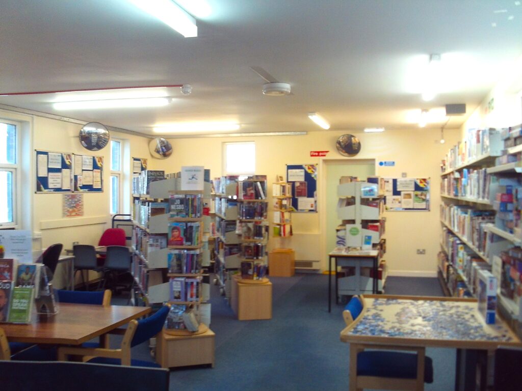 Photograph of the north site library, a room in which there are book shelves of various styles, some tables, and information displays on the walls. The shelves are all well stocked with books.
