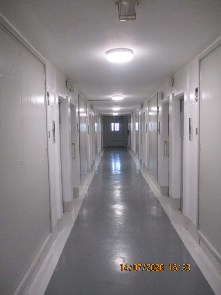 Photograph of a communal corridor, with doors leading off to both sides and a window at the far end leting light in. It is obvious that the space is very clean - there are no signs of grime and the vinyl floor reflects light.