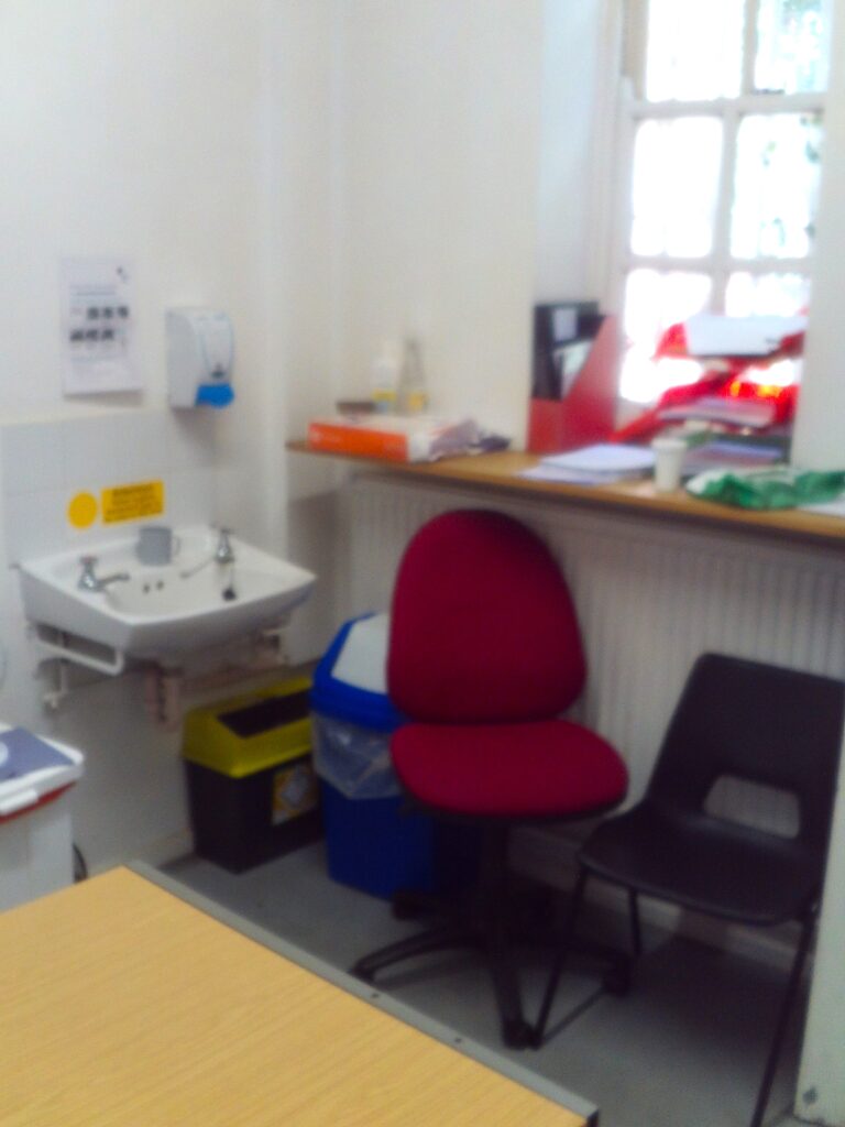 Photograph of a health room in the reception area. A desk, chair, sink, bins and folders can be seen.