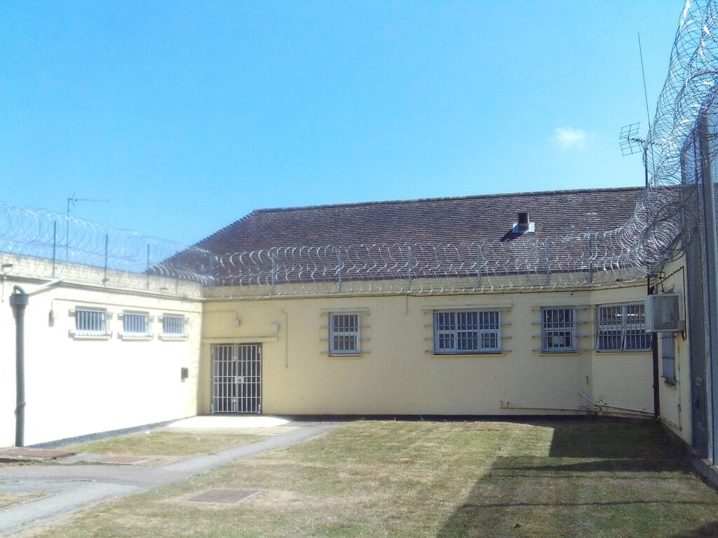 Photograph of the patient entrance to the health building. The one-story building is painted a cream colour and has windows in its walls. Barbed wire is arranged across the guttering.