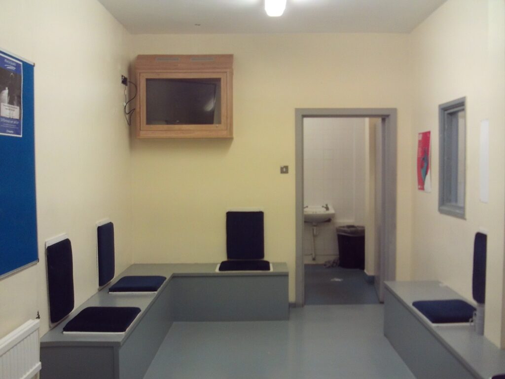 Photograph of the reception holding room. Wooden benches are set against three of the walls and have pads on them for people to sit on; the walls are painted cream and there is a noticeboard mounted on one wall.