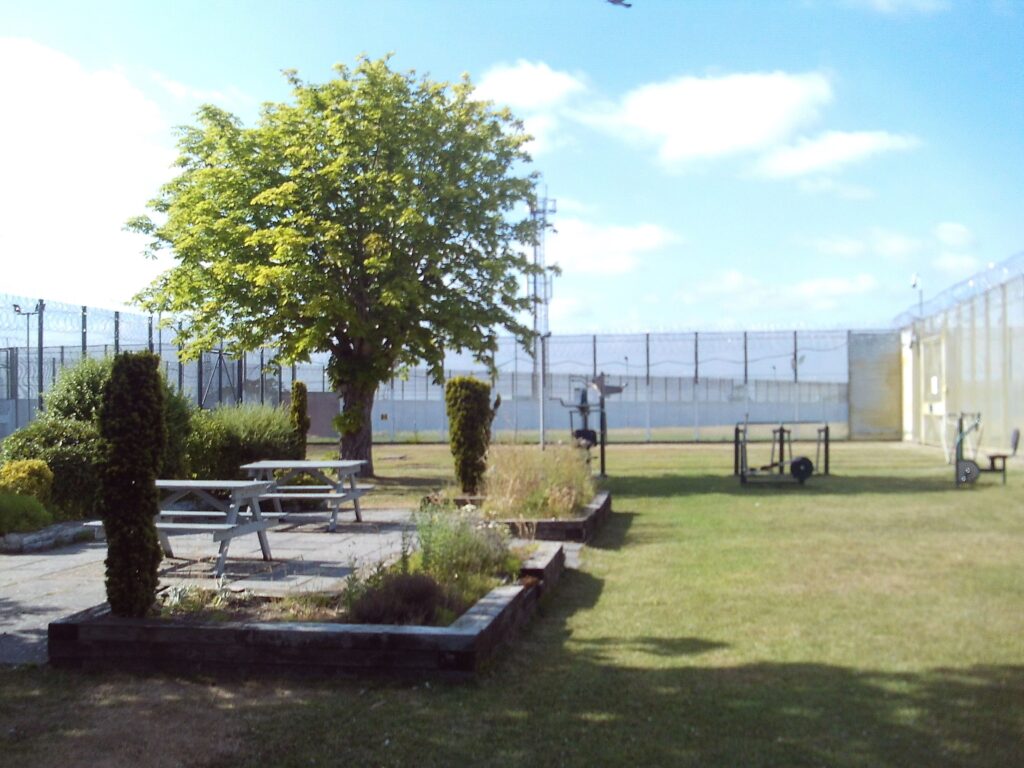 A photograph of the ISFL unit garden. There are trees and shrubs, a grass area, some picnic tables and some exercise equipment. There is a high fence enclosing the area.