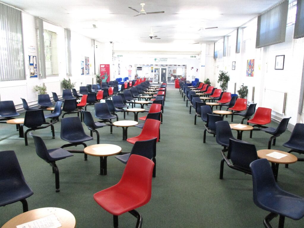 A photograph of the interior of the Visits Hall on the south site. Fixed tables and chairs are arranged in rows down the length of the room.