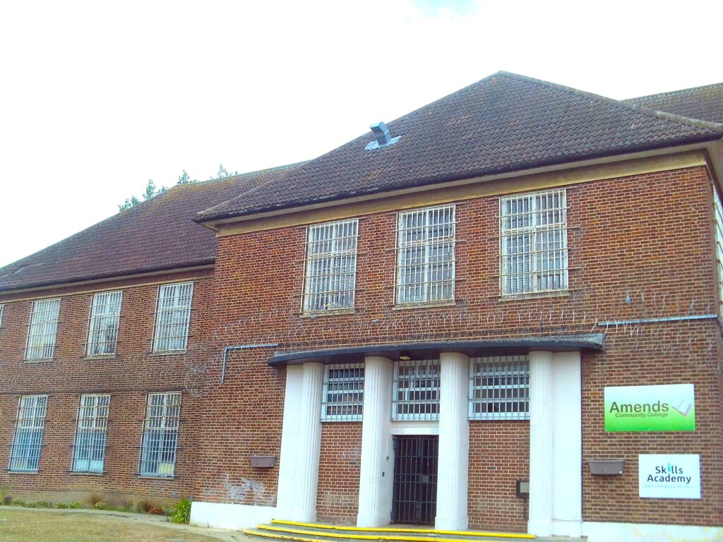Photograph of the exterior of one of the buildings at HMP Highpoint. It is a two-storey, red brick building with with windows on both floors and a 'Skills Academy' sign on one side.