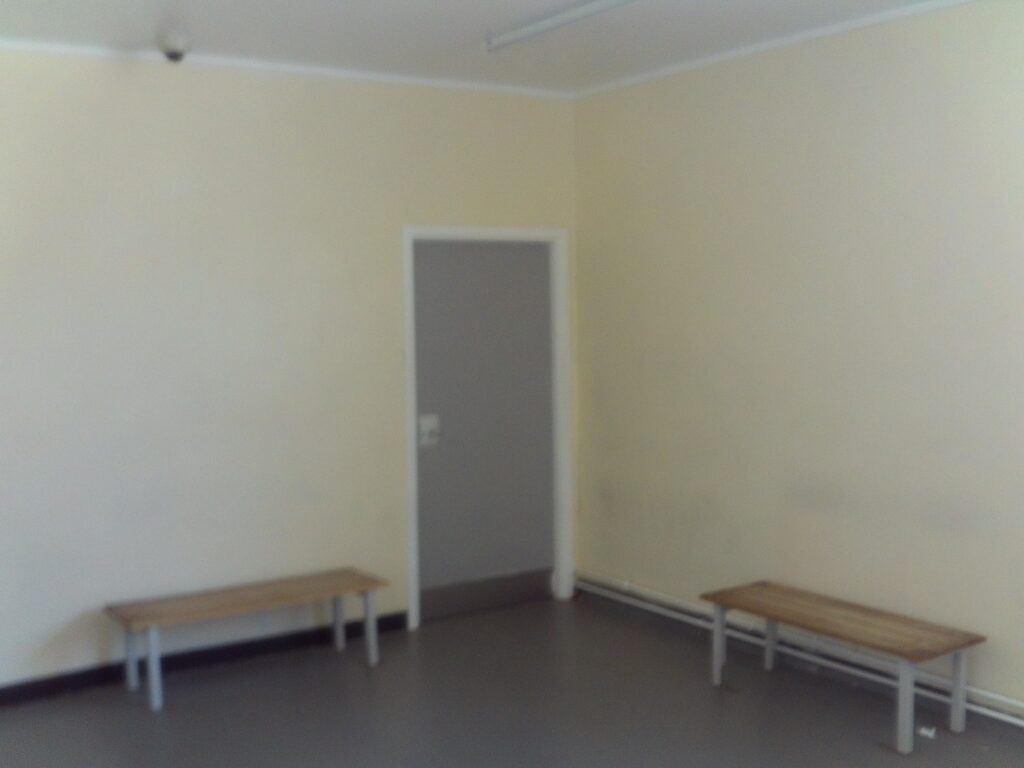 Photograph of a patient waiting area, a room with the walls painted yellow and a grey door in one corner. Benches are positioned against two walls.