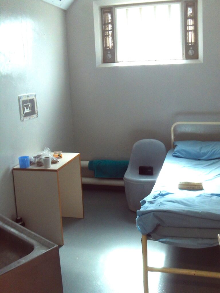 Photograph of a segregation unit cell. There is a bed to one side, with a plastic chair next to it. A table can be seen against the opposite wall.