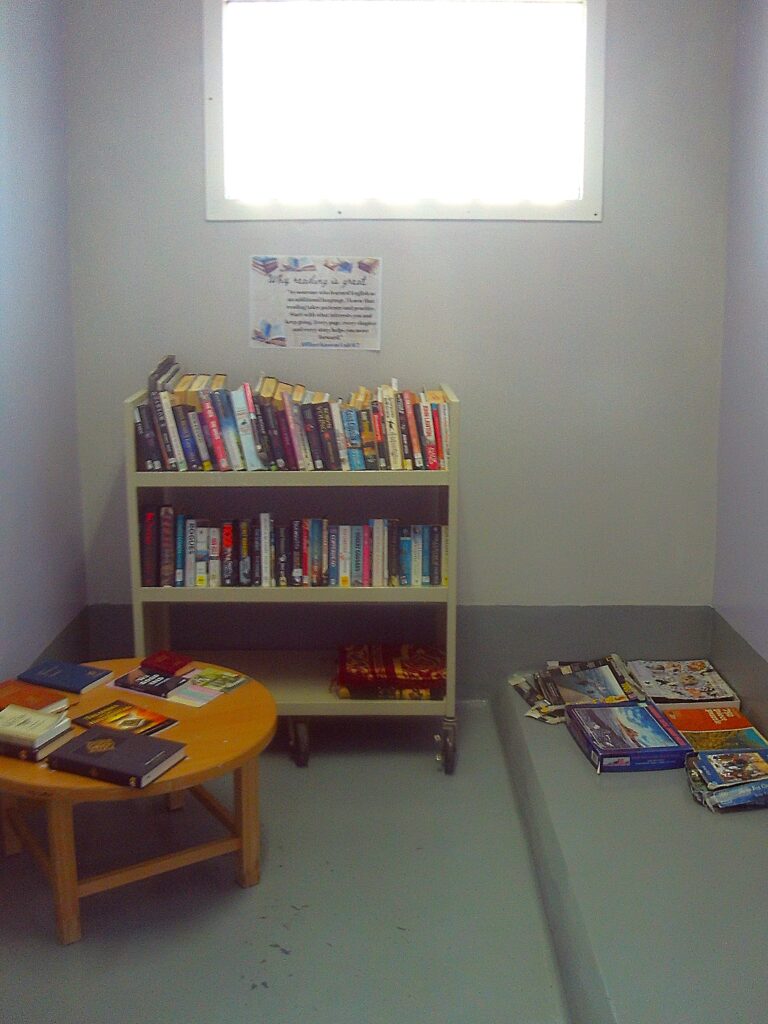 Photograph of the segregation unit library, a small room with a book shelf stocked with books, some board games and a coffee table in it.