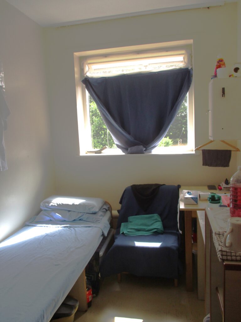 Photograph of a cell on B wing. A bed is one side of the room, with a desk and other furniture on the other side. A chair is between them, under the window.