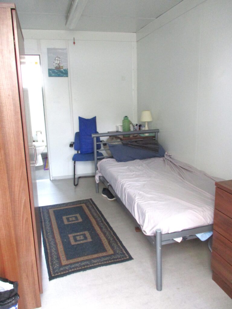 Photograph of a C$ pod room. there is a bed on one side, along with some drawers, there is a rug on the floor and a wardrobe beside it.