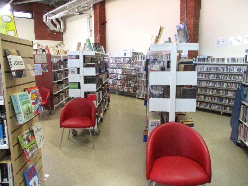 Photograph of the interior of the library, many shelves of books can be seen, and there are some chairs positioned at the end of some of them.